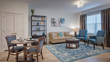 Living room with white walls, light wood floor, tan couch with two blue chairs, round ottoman table over blue rug, dark wood bookshelf to left of couch and small round table with two chairs to left of room.