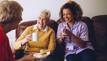 Senior woman sitting on sofa and holding a cup of coffee next to a caregiver holding a coffee cup and across from another senior woman holding a plate with coffee cake.