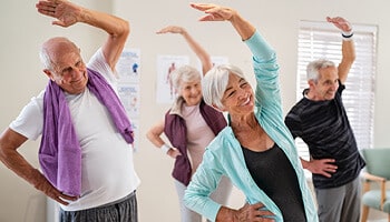 Two senior couples wearing exercise clothes standing and stretching their left hands over their head with right hands on hips.