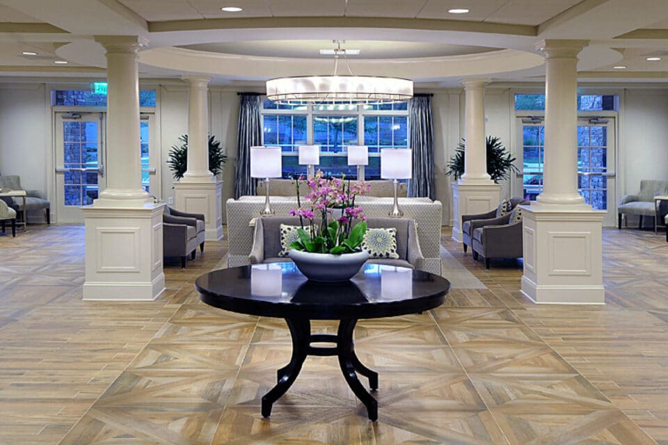 Lobby area with white columns, dark wood table at center and floral centerpiece, gray chairs and patterned sofas at center, with wood floor.