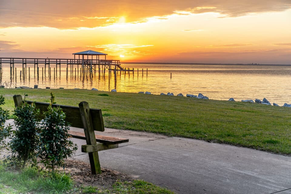 View of sunset on the bay with a pier at left and an empty park bench.
