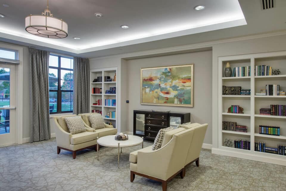 Living room with bookcases on each side of the back wall, four beige sitting chairs at center of room with round white table in the middle and windows to the left of room.