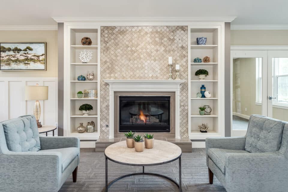 View of living room of with fireplace at back wall with tile that goes to ceiling, white bookcases on each side, two blue sitting chairs in center of room with round table in the middle and an end table at left with a lamp.