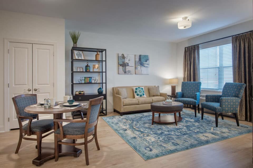 Living room with white walls, light wood floor, tan couch with two blue chairs, round ottoman table over blue rug, dark wood bookshelf to left of couch and small round table with two chairs to left of room.