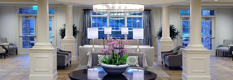 Lobby area with white columns, dark wood table at center and floral centerpiece, gray chairs and patterned sofas at center, with wood floor.
