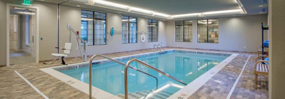 Indoor swimming pool with brown tiled floor and windows at back of room.