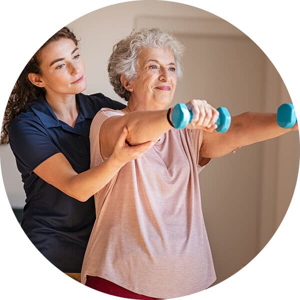 Senior woman raising hand weights in front of her with caregiver standing at left and offering support.