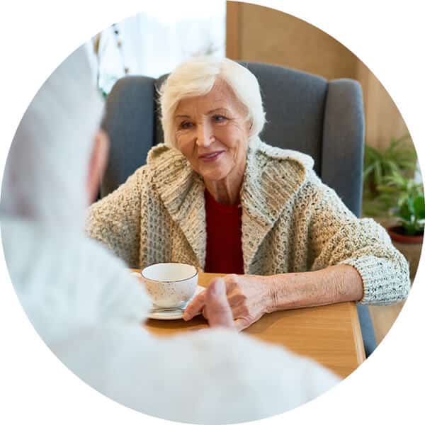 Senior woman sitting in blue chair at table and enjoying coffee and conversation with another senior woman.